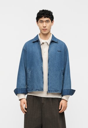 Ung mand iført en blå denimjakke over en beige skjorte med krave og mørkebrune bukser, stående foran en ensfarvet baggrund.