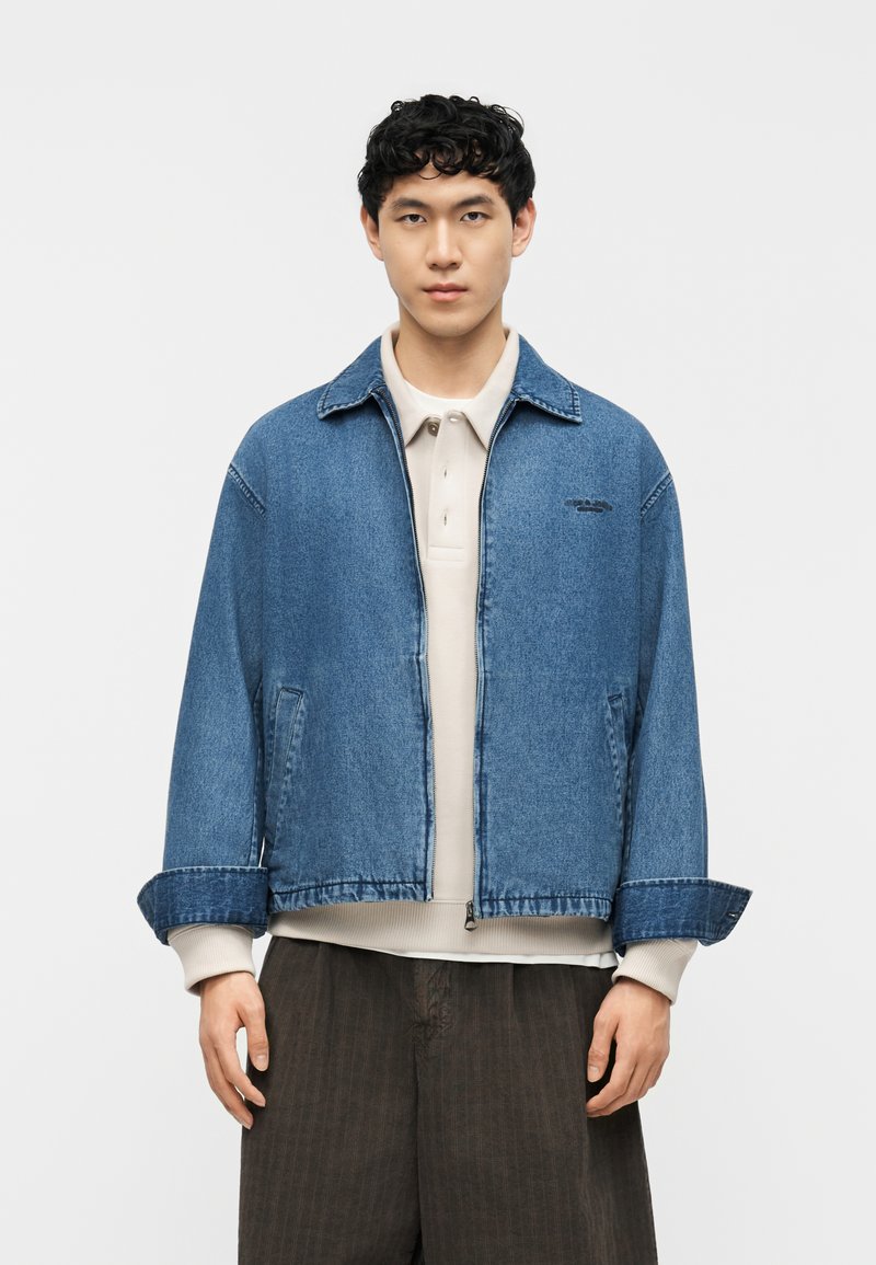 Νέος άνδρας που φοράει μπλε τζιν μπουφάν πάνω από μια μπεζ πουκαμίσα με γιακά και σκούρο καφέ παντελόνι, στέκεται μπροστά σε ένα απλό φόντο.