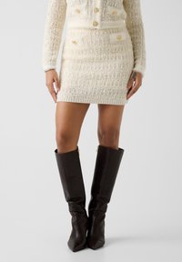 Robe pull deux pièces en maille crème avec détails de boutons dorés, tissu texturé et manches longues, associée à des bottes en cuir marron montant jusqu'aux genoux.