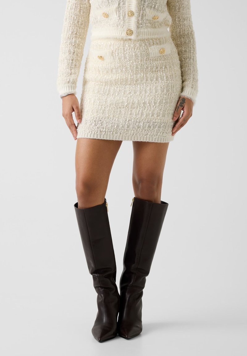 Robe pull deux pièces en maille crème avec détails de boutons dorés, tissu texturé et manches longues, associée à des bottes en cuir marron montant jusqu'aux genoux.