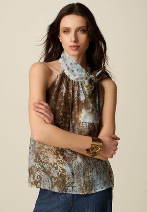 Jeune femme portant un haut sans manches à motif cachemire et floral, avec un nœud et un bracelet manchette doré, debout les bras croisés.