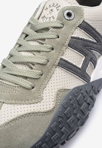 Baskets en suède beige et verte avec des détails en maille, présentant un logo accent gris et une semelle noire texturée. Les lacets sont dans un vert coordonné.