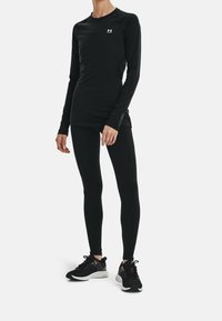 Czarny, długi rękaw, sportowa bluzka z rękawami raglanowymi, w połączeniu z czarnymi legginsami i czarnymi butami sportowymi z szarymi akcentami. Widoczne logo Under Armour.