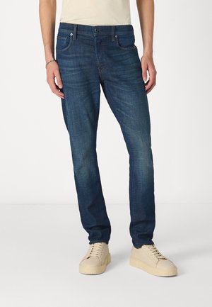 Dark blue denim jeans with a straight-leg cut, five-pocket design, and slight fading. Worn with beige sneakers.