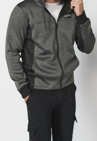 Veste grise à fermeture éclair avec des accents noirs, en tissu texturé ; comprend une capuche, des cordons de serrage et des poches latérales. Associée à un pantalon noir.