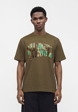 Jeune homme portant un t-shirt vert olive avec un texte coloré "Billionaire Boys Club" et un short noir, debout devant un fond blanc uni.