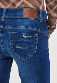 Mėlyni džinsai iš džinso audinio su odinėmis etiketėmis, pažymėtomis užrašu „Pepe Jeans“, su geltona siūle ir galine kišene su siuvinėtu logotipu.