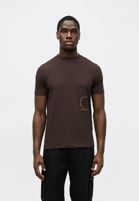 T-shirt de algodão castanho com mangas curtas, apresentando um design em contorno dourado na área esquerda do peito. Ajuste clássico, textura suave.