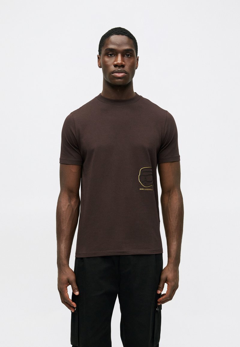 T-shirt de algodão castanho com mangas curtas, apresentando um design em contorno dourado na área esquerda do peito. Ajuste clássico, textura suave.