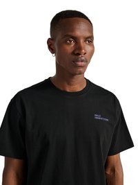 Homme aux cheveux courts et boucle d’oreille en forme d’anneau, portant un t-shirt noir avec un petit texte blanc "HALO 19681972981" sur la poitrine, regardant vers la droite.