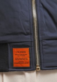 Veste de pilote bomber bleu marine avec fermeture éclair argentée et étiquette orange cousue indiquant les spécifications militaires et les détails de propriété.