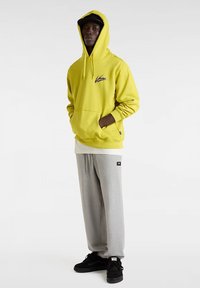 Felpa gialla con tasca frontale e logo, abbinata a pantaloni della tuta grigi e sneakers nere. Il materiale sembra morbido con una vestibilità comoda.