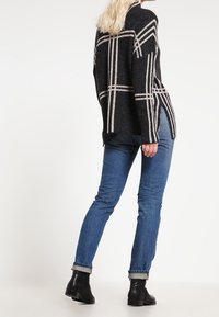 Czarny i beżowy sweter w kratę z bocznymi rozcięciami, w zestawieniu z obcisłymi niebieskimi dżinsami i czarnymi botkami. Miękka tekstura, casualowy fason.