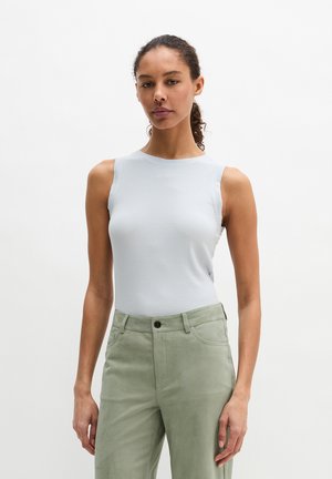 Femme debout devant un fond blanc, portant un haut blanc sans manches et un pantalon taille haute vert clair, regardant devant elle avec une expression neutre.