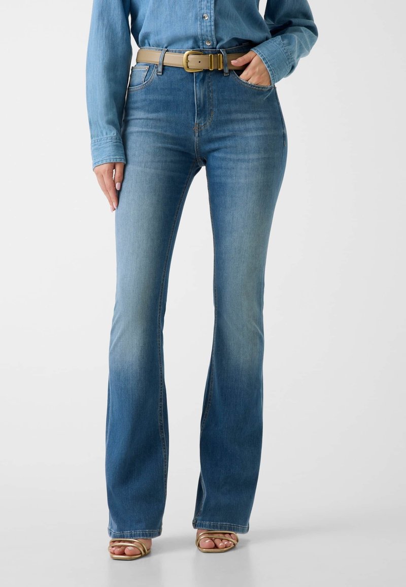 Femme portant un jean bootcut bleu délavé, ceinture beige, chemise en denim et sandales dorées à bout ouvert, debout avec une main dans la poche.