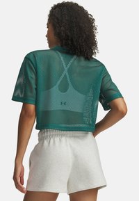 Krotka majica iz turkiznega mrežastega materiala z okroglim izrezom in kratkimi rokavi, slojevito oblečena čez svetlo barvni športni nedrček. Kombinirano s svetlo sivimi kratkimi hlačami.