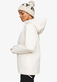 Helle, wasserdichte Jacke mit Kapuze, kontrastierender Brusttasche und verstellbaren Manschetten. Kombiniert mit einer Strickmütze mit schwarzen Akzenten.