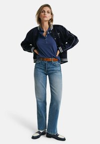 Marine bomberjack met witte strepen, blauwe polo, hoog-wijd lichtblauwe denim jeans, bruine riem en twee-kleurige loafers.
