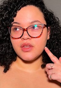 Person med krøllete hår som har på seg store, skjellsølje-briller, peker på kinnet sitt. Lys hudtone, leppene er litt åpne, ringer på fingrene.