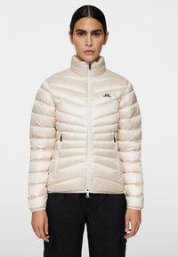 Beżowa kurtka puchowa z poziomymi przeszyciami, wysokim kołnierzem, z przodu na zamek oraz dwiema kieszeniami po bokach. Zawiera logo na lewym piersi i ma gładką teksturę.