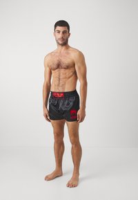 Schwarze Satin-Boxershorts mit rotem "VENUM"-Logo und Akzenten, ausgestattet mit einem elastischen Bund und einer lockeren Passform. Das Modell ist barfuß.