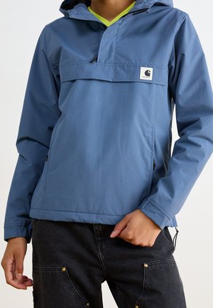 Personne portant une veste coupe-vent bleue à capuche sur une chemise vert citron, associée à un pantalon noir avec des poches avant et des rivets dorés.