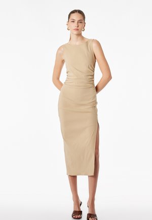 Femme portant une robe midi beige sans manches avec une fente latérale, des talons marron ouverts à l'avant et des boucles d'oreilles créoles dorées, debout devant un fond blanc.