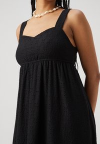 Černé šaty z texturované látky se čtvercovým výstřihem, širokými ramínky a řaseným pasem, s vystřiženými vzory po celém povrchu.