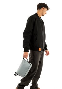 Bomber nero con polsini a costine, pantaloni grigi e una borsa grigio chiaro con texture, tenuta per il manico, con cerniera nera e dettagli in nero.