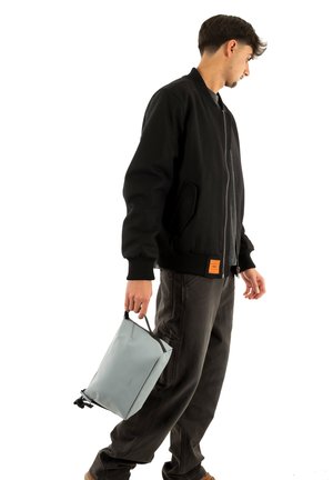 Bomber nero con polsini a costine, pantaloni grigi e una borsa grigio chiaro con texture, tenuta per il manico, con cerniera nera e dettagli in nero.