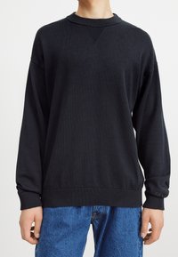 Schwarzes Sweatshirt mit einem gerippten Ausschnitt und strukturierten Details, mit überschnittenen Schultern und einem umsäumten Saum. Kombiniert mit blauen Jeans.