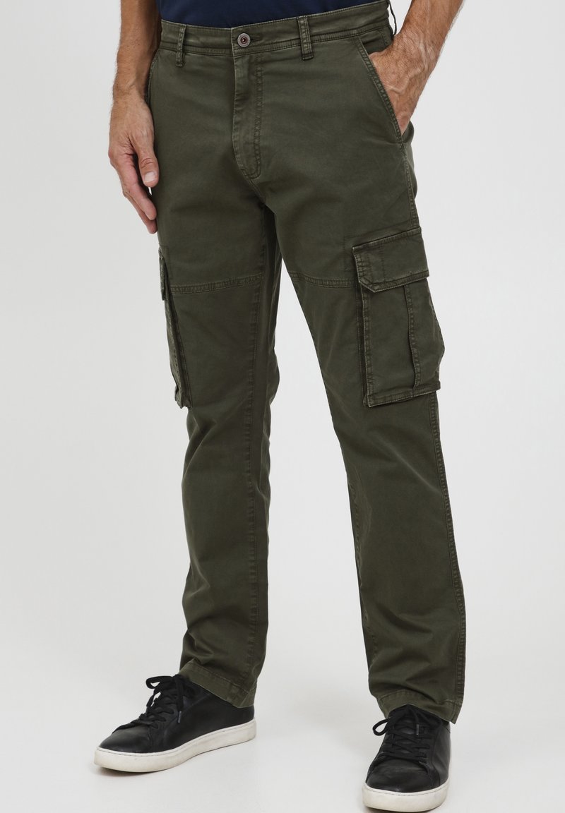 Homme portant un pantalon cargo vert olive avec plusieurs poches et des baskets noires, debout les mains partiellement dans les poches.