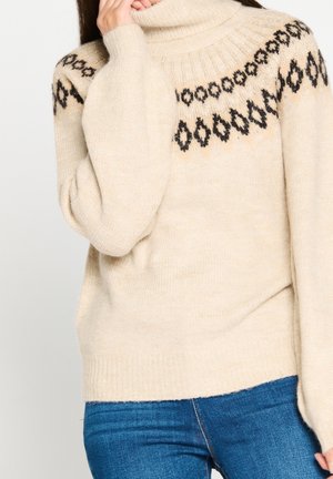 Pull à col roulé beige avec motif géométrique noir sur le yoke, porté avec un jean bleu, personne tenant le col près du visage.