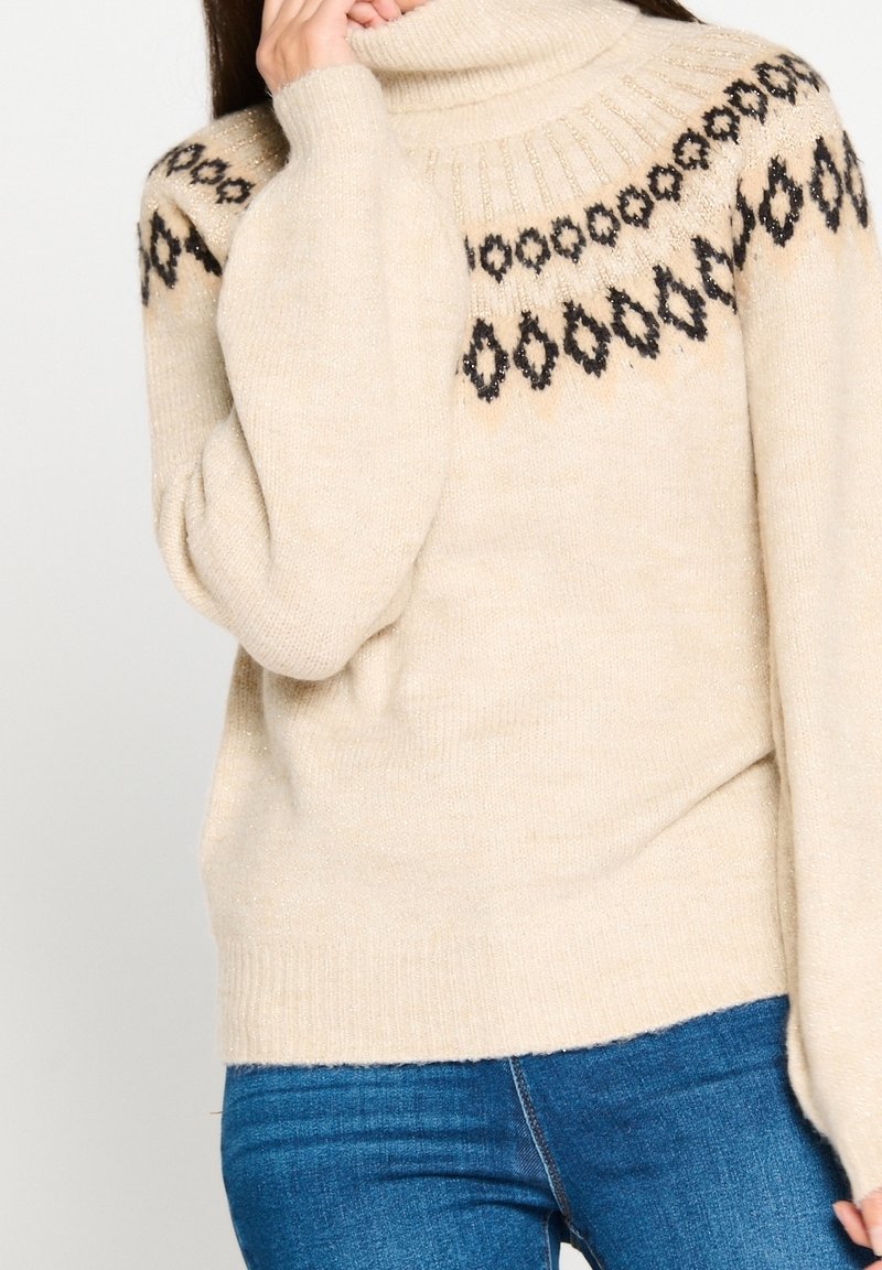 Pull à col roulé beige avec motif géométrique noir sur le yoke, porté avec un jean bleu, personne tenant le col près du visage.