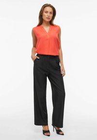 Blusa rossa senza maniche con scollo a V, abbinata a pantaloni larghi neri con tasche laterali e una piega davanti.