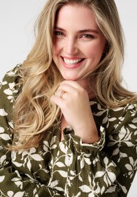 Blouse vert olive clair avec des motifs de feuilles blanches, tissu texturé, manches bouffantes, et une bague en or superposée au doigt. Sourire confiant.