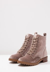 Bottines en daim marron avec un design à lacets, bout arrondi et talon bloc de 3 cm. Elles disposent d'une semelle en caoutchouc et d'œillets en métal sur les lacets.