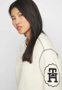 Maglione bianco testurizzato con accenti neri e toppa con logo ricamato sulla manica. Presenta cuciture a contrasto e una vestibilità morbida.
