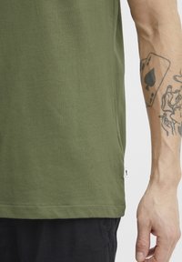 Olivengrønn bomull T-skjorte med rett snitt, korte ermer og en liten logoetikett nederst; glatt tekstur og minimalistisk design.