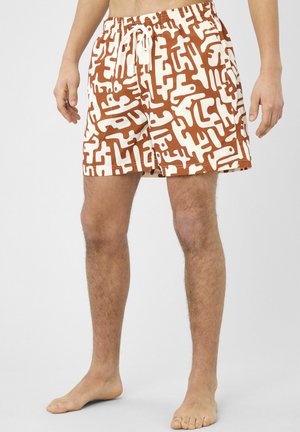 Jambes d'homme portant un short de bain à motif abstrait marron et blanc avec un cordon blanc, debout pieds nus sur un fond uni.
