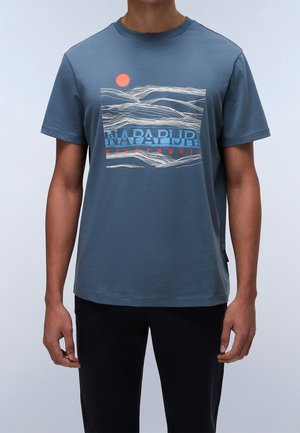 Mand iført blå kortærmet t-shirt med hvid linjekunst af bjerge, rød sol og blå og rød "Napapijri Geographic" tekst på forsiden.