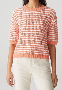 Mujer vistiendo un suéter de manga corta texturizado con franjas horizontales en coral y blanco, y pantalones de color claro, frente a un fondo sencillo.