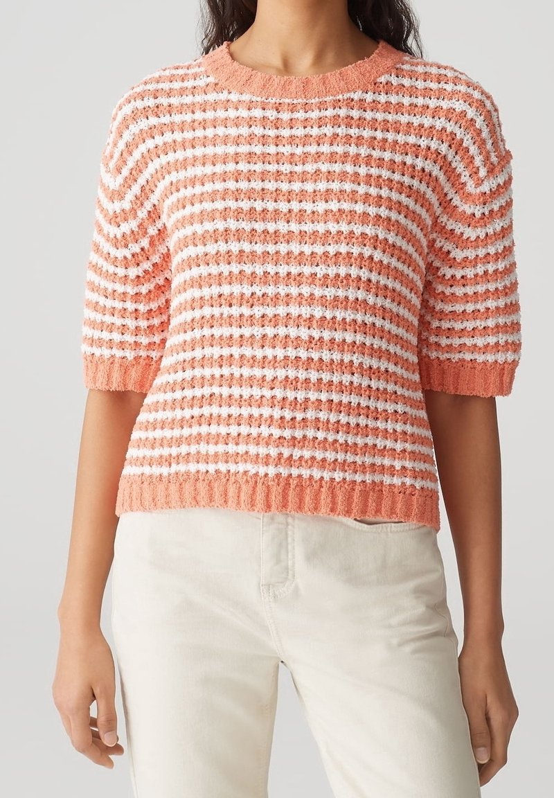 Mujer vistiendo un suéter de manga corta texturizado con franjas horizontales en coral y blanco, y pantalones de color claro, frente a un fondo sencillo.