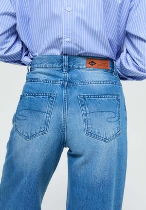Des jeans en denim bleu taille haute présentent un délavage, deux poches arrière avec des détails de couture et une étiquette marron au niveau de la ceinture.