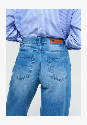 Des jeans en denim bleu taille haute présentent un délavage, deux poches arrière avec des détails de couture et une étiquette marron au niveau de la ceinture.