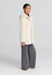 Cappotto in finta pelliccia color crema con cappuccio, maniche lunghe e bottoni frontali. Abbinato a pantaloni grigi larghi e scarpe slip-on color crema.