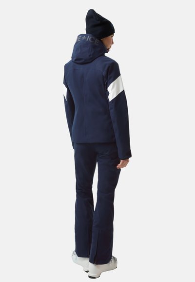 Granatowa kurtka narciarska z białymi akcentami i logo na kołnierzu, w komplecie z pasującymi spodniami. Posiada kaptur oraz zamki błyskawiczne w okolicy kostek.