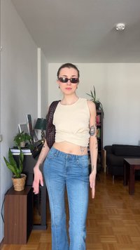 Moteris su akiniais nuo saulės, džemperiu be rankovių kremine spalva ir mėlynomis džinsais stovi svetainėje, kur matyti augalai, knygos ir tamsi sofa fone.