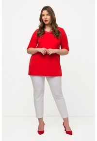 Rode tuniektop met korte mouwen, versierd rond de halslijn. Gekozen in combinatie met witte enkellange broeken en rode hakken.