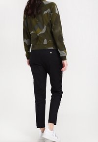 Un pull vert olive avec des formes abstraites grises et jaunes, associé à un pantalon noir ajusté et des baskets blanches. Texture lisse partout.